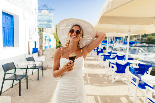 Gorgeous young woman eating ice cream and walking on the street at summer in the beautiful village Fiscardo on Kefalonia island in Greece. Beautiful woman wearing a hat, sunglasses and white dress, has a long blond hair and looks very happy