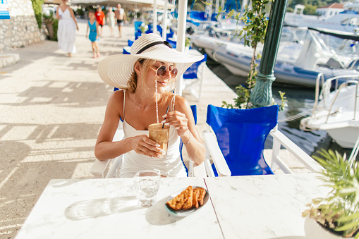 Gorgeous young woman relaxing in restaurant near marina with yachts and boats on sea and enjoying while drinking morning coffee at sunny day. She is on vacation and looks happy and carefree