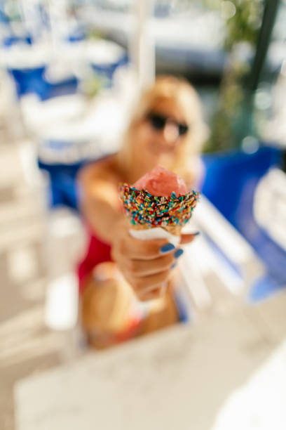 Gorgeous young woman relaxing in restaurant near marina by the sea and enjoying while eating refreshing ice cream at sunny day. She is holding ice cream toward camera