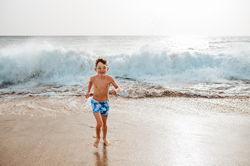 Young boy playing, running and splashing in strong sea waves. Smilling boy in swimsuit and swimming goggles. Concept of a beach summer vacation with kids.
