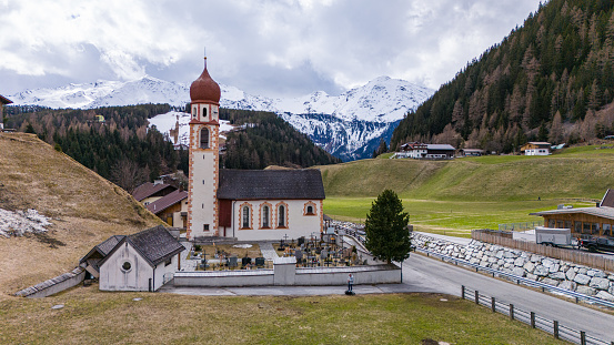 Aerial drone view of Niederthai in Austria. Beautiful onion dome church.  Early springtime view, with snowcapped mountains in the background. Grassy slopes and pine trees.