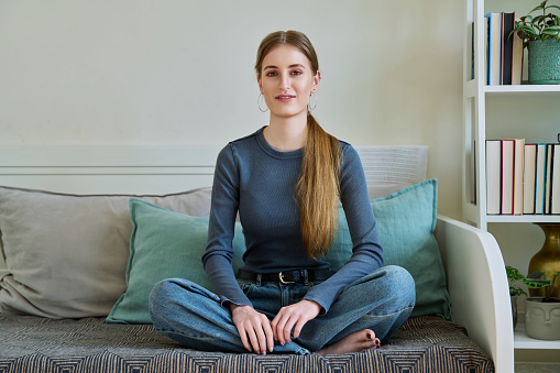 Portrait of happy attractive 17,18 year old relaxed teenage girl sitting in lotus position on couch at home, smiling looking at camera. Youth, lifestyle concept