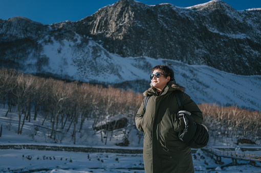 Asian Chinese mature female tourist standing in front of snowcapped mountain looking away admiring scenery
