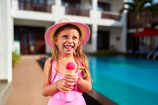 Child relishes sweet treat summer sun, water reflections create luminous ambiance. Happy girl in pink swimsuit enjoys strawberry ice cream by blue poolside. Leisure, innocence, childhood pool moment.