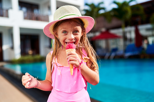 Child with delightful expression savors summer treat, vacation ambiance, warm sunny day, leisure time. Little girl in pink swimwear and sun hat enjoys strawberry ice cream by bright blue poolside.