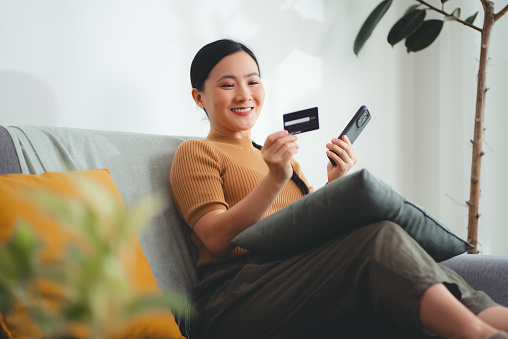Asian woman sitting on sofa in living room using smart phone and credit card for shopping online at home. Happy excited woman shopper using instant mobile payments making purchase in online store.