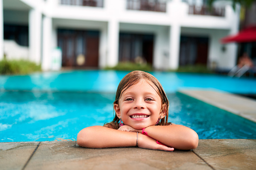 Child with wet hair leans on edge, happy in water. Smiling girl enjoys summer fun in resort pool. Bright day for kids outdoor swimming activity. Playful moment, family vacation joy at tropical hotel.