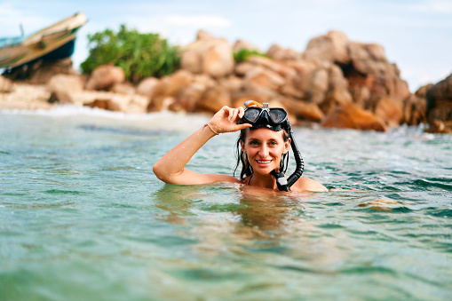 Female enjoying snorkeling adventure in sea near beach. Smiling woman swims in clear ocean by rocky coast, adjusts diving goggles. Leisure activity in tropical water. Holiday swim, exploration.