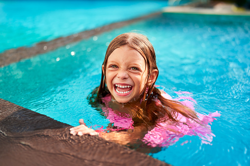 Sunlit water sparkles around joyful child enjoying summer vacation. Happy girl in pink swimsuit swims, plays in kid-friendly resort pool. Family-friendly leisure, splash fun, tropical getaway scene.