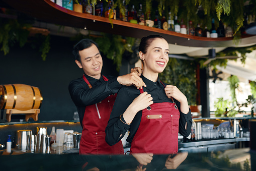 Friendly bartender helping coworker to adjust apron strap