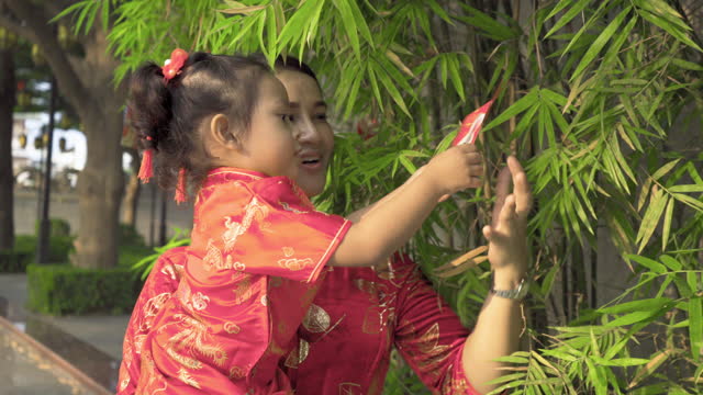 Chinese mother and daughter dressed in red Chinese cheongsams, looking at red cards hung on bamboo trees.