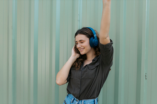 Attractive young woman standing against metal wall and listening to music on headphones. Pretty female model in casuals enjoying listening to music on wireless headphones.