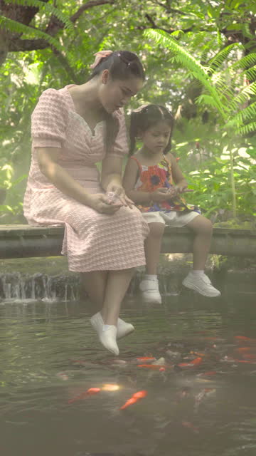 Asian mother and daughter feeding koi fish in a park with mist and shady nature.