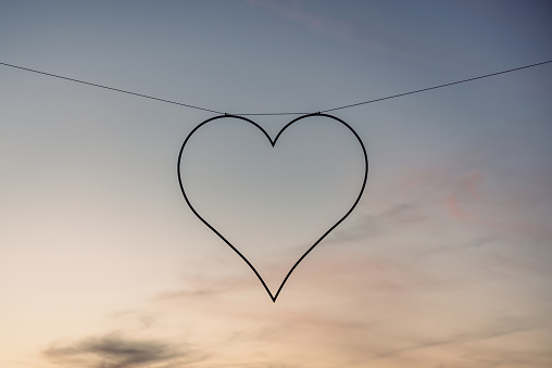 A delicate heart-shaped silhouette dangles from a thin wire, suspended against a soft gradient of twilight hues transitioning from day to night. The simplicity of the heart contrasts with the complexity of the skys natural palette.