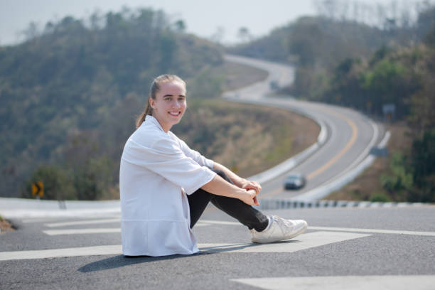 Young blond woman with long hair in retro orange jumpsuit is on the country empty highway traveling in summer.