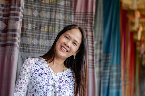 Asian mid adult woman in white t-shirt holding woven fabric which was decorated on ceiling of local cafe for tourist watching and taking photos, soft focus, happiness of humans concept.
