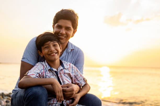 Warm outdoor portrait of a father hugging his son, with a serene sunset over the sea in the background, depicting family love and bonding.