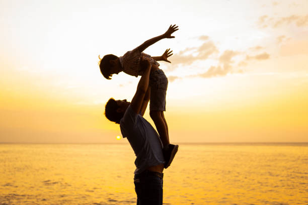 Silhouetted father joyfully lifting his child in the air on a serene beach as the sun sets, depicting family fun and bonding.