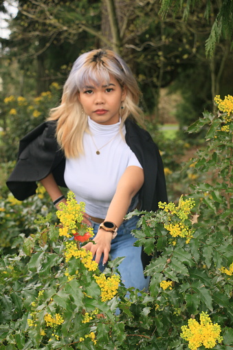 A Filipino woman with yellow flowers in  a public garden in Spring. She is wearing medium length, multi colored hair, a white tee shirt, necklace, black jacket, jeans, a watch and bracelets.