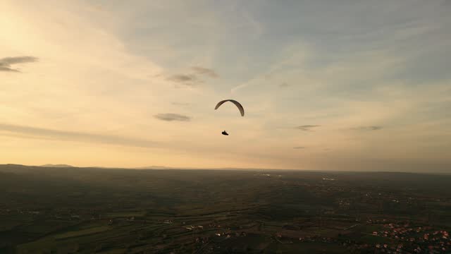Motorized paraglider flies low over 
 rural land in sunset.