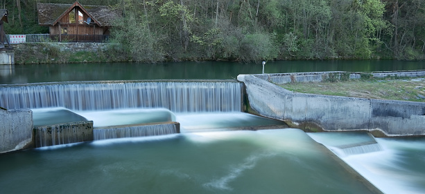 This serene image is a time-blended capture from a 4k video, showcasing the smooth, flowing beauty of small cataracts in the Loisach at Wolfratshausen. The technique averages the visual data over time, resulting in a silky portrayal of the river's cascading waters. The ethereal tranquility of the scene, combined with the detailed texture of the concrete structures and the lush green backdrop, creates a harmonious blend of human engineering and nature's artistry, perfect for a range of uses from environmental studies to artistic displays.