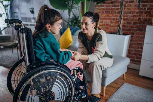 A mother sits beside her daughter who is in a wheelchair, their hands lovingly connected. The warmth of their bond illuminates the cozy, well-decorated living space around them