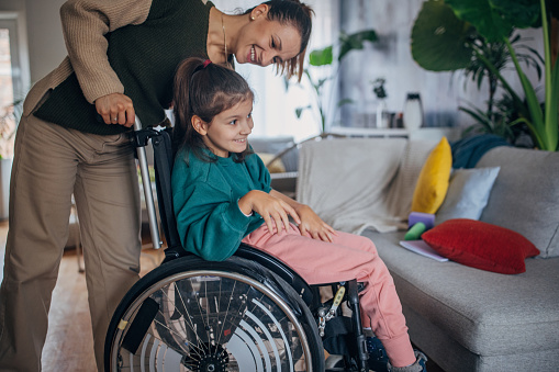 A heartwarming scene unfolds in a cozy living room where a mother and daughter share a playful moment. The daughter, seated in a wheelchair giggles , their smiles illuminating the space with warmth and love.