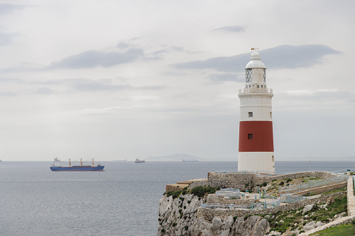 A lighthouse is on a rocky cliff overlooking the ocean. The lighthouse is red and white. A large ship is in the distance