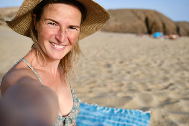 Smiling young female in summer hat looking at camera while standing on blanket in sunlight and taking selfie on sandy beach with blurred tourists near rocky hills