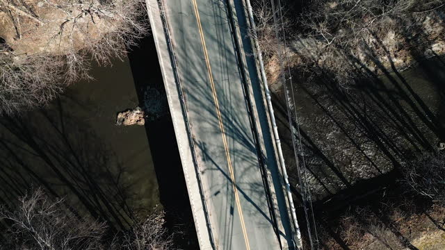 Drone ascends above calm empty bridge as sedan and SUV drive across asphalt, long tree shadows