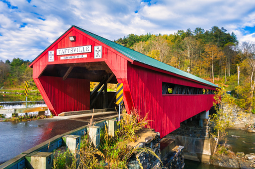 Woodstock, Vermont, USA- October 12, 2023-  A momentary break in the clouds after a rain shower lets a bit of sun hit  the red Taftsville Covered Bridge,  a timber-framed covered bridge which spans the Ottauquechee River in the Taftsville village of Woodstock, Vermont.