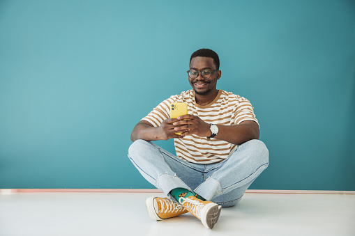 Portrait of young black man on blue background, using smart phone.