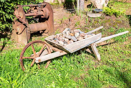 Old wooden cart with a big metal wheel and a bunch of stones in summer garden