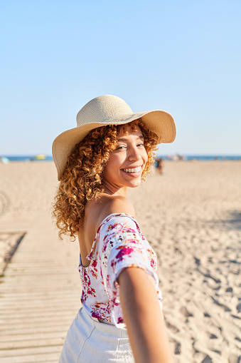 Smiling young woman with braided hair holding hands on the beach. Dressed in a straw hat and casual clothes. Concept of happiness