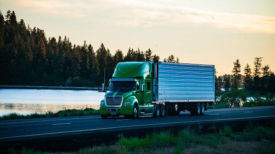 A green long-haul semi-truck with a refrigerated trailer, driving along a scenic section of interstate freeway just after sunrise, with trees and a lake in the background.