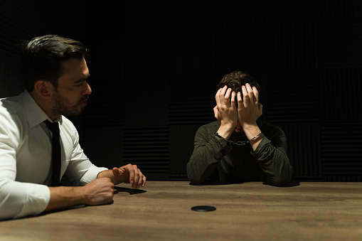Lawyer talking to a handcuffed suspect showing signs of distress in a dimly lit interrogation room