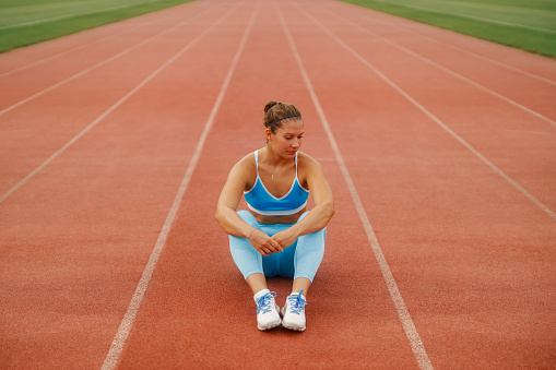 A fit sportswoman in shape is sitting on running track at stadium and resting form exercises. A tired female runner in shape is sitting ground at stadium, taking a break and resting form running.