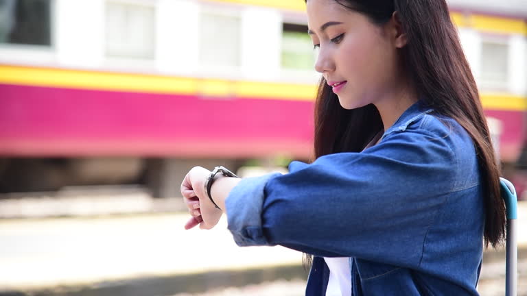Asian young woman traveler backpack waiting train at railway station look at hands watch. Women holding ticket passenger passport on hot sunlight day. Stress Female tourist travel transportation trip