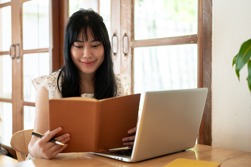 A beautiful young Asian woman is focusing on reading a book, working remotely at a coffee shop. people and lifestyle concepts