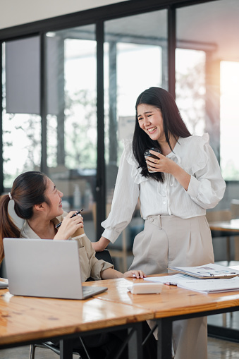 Two professional women sharing a light-hearted moment and a laugh over a coffee break in a bright, modern office setting.