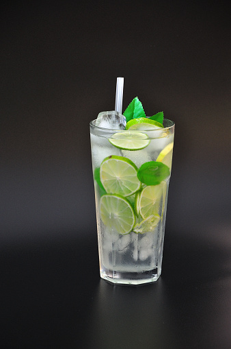 Tall faceted glass of refreshing lemonade with ice and mint on a black background, next to pieces of ripe lime. Vertical arrangement.