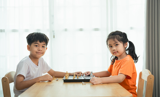 Two children are playing a game of chess. One of the children is wearing an orange shirt. The children are sitting at a wooden table