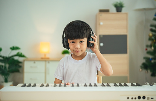 A young child wearing headphones is sitting at a keyboard. The child is wearing a white shirt and he is focused on playing the keyboard