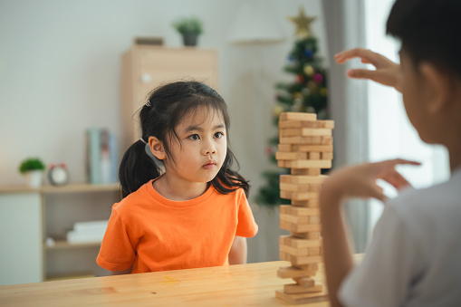 A girl in an orange shirt is sitting at a table with a stack of wooden blocks. A boy is standing behind her, watching her. Scene is playful and lighthearted