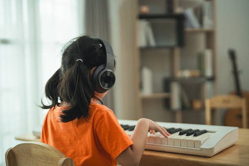 A young girl is sitting at a piano, wearing headphones. She is wearing an orange shirt. The room is filled with books and a keyboard