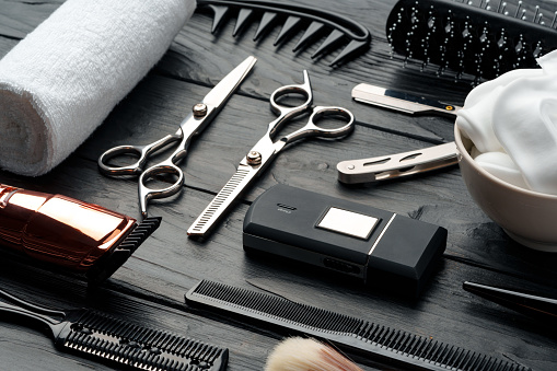 A variety of professional barber tools are neatly arranged on a dark wooden surface, including scissors, a straight razor, combs, clippers, and grooming products, suggesting preparation for a grooming session.