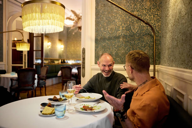Mature men talking and flirting during dinner date Casually dressed couple seated at table for two in elegant restaurant, interacting and laughing as they enjoy their meal and white wine. gay dinner table stock pictures, royalty-free photos & images