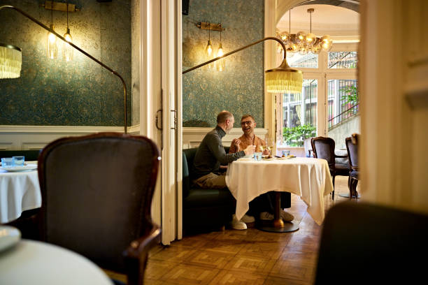 Mature men talking and flirting during lunch date Focus on background table for two with gay couple smiling, talking, and laughing while enjoying midday meal and wine. gay dinner table stock pictures, royalty-free photos & images