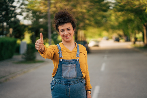 Portrait of a smiling woman with a curly hairstyle standing on the street and showing thumbs up.