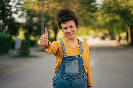 Portrait of a smiling woman with a curly hairstyle standing on the street and showing thumbs up.